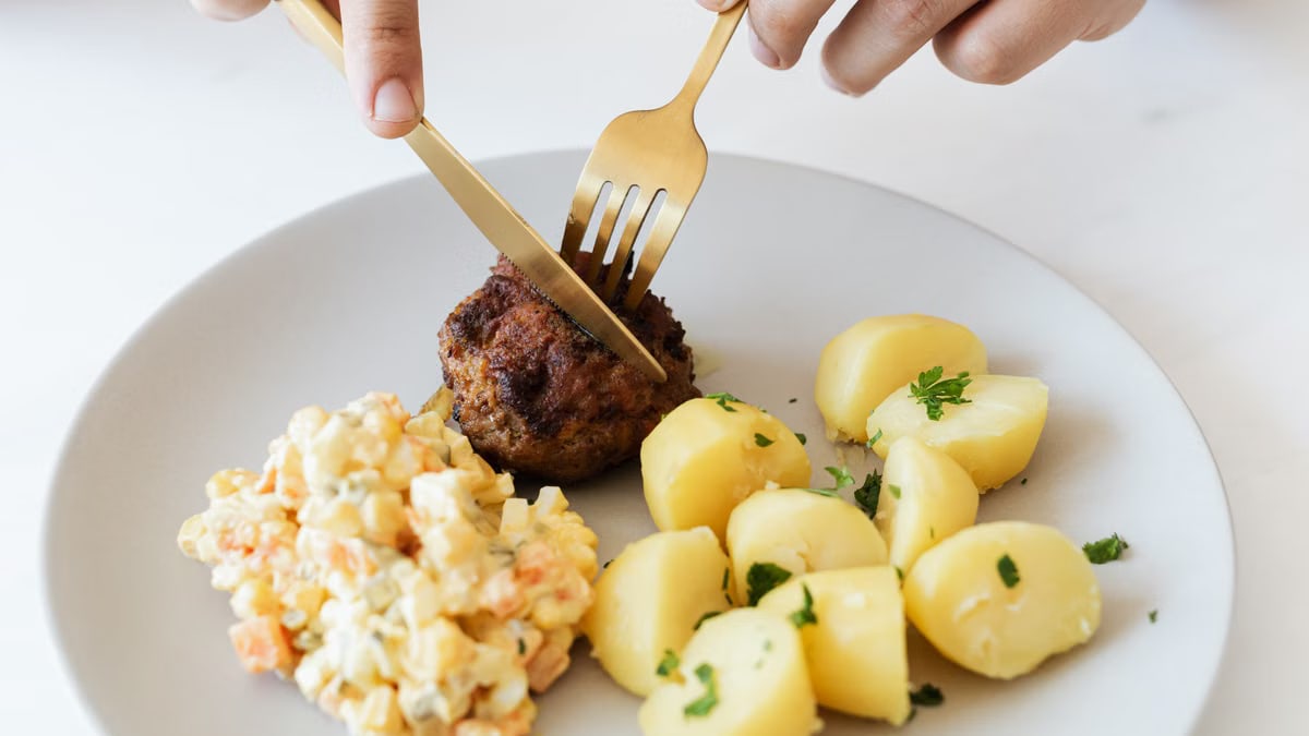From above of crop anonymous person cutting with cutleries fried juicy meat cutlet served on white plate with tasty boiled potatoes and Russian salad